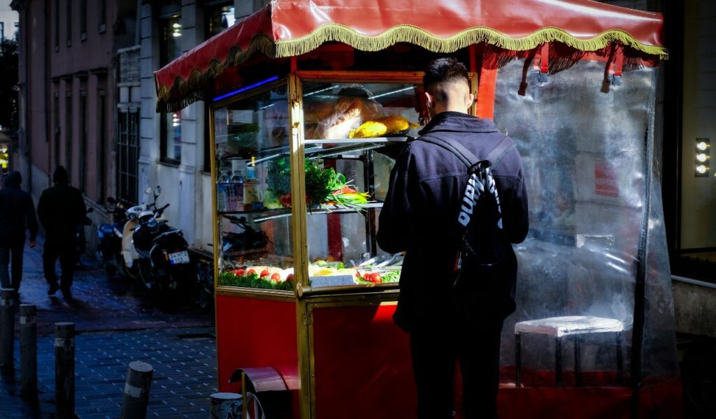 A man standing at a brightly lit street food cart in Istanbul, with fresh produce and snacks displayed inside the glass case at night.