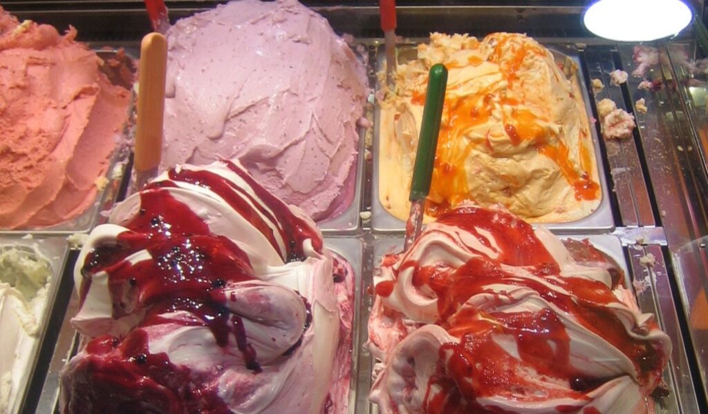 Colorful trays of Italian gelato in various fruity flavors with swirls of syrup, displayed at a street-side gelateria.