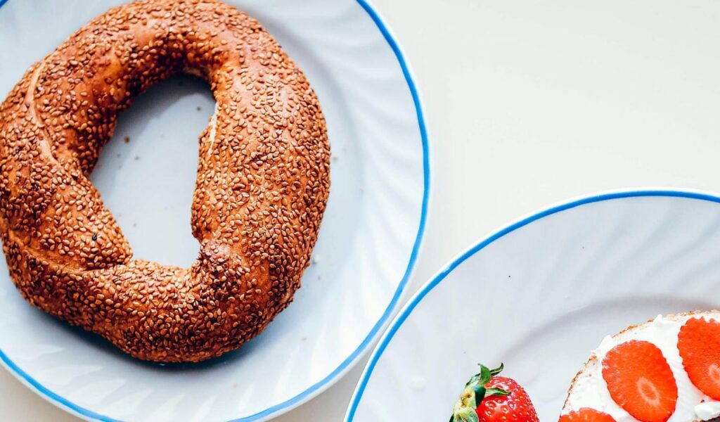 A sesame-coated Jerusalem bagel sits on a plate next to another plate with cream cheese bagel slices topped with strawberries and fresh strawberries on the side.