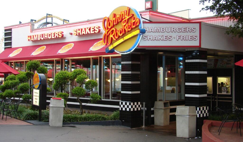 Exterior of Johnny Rockets diner with red awnings and neon signage for burgers and shakes.