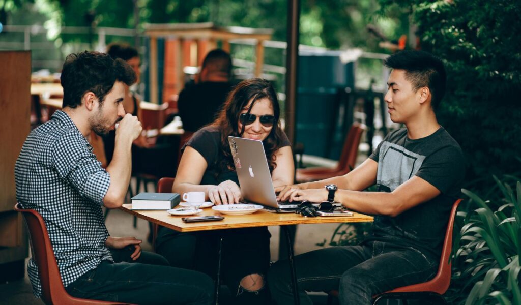 Three young adults sitting at an outdoor café table, working together on a laptop and having coffee, surrounded by greenery.