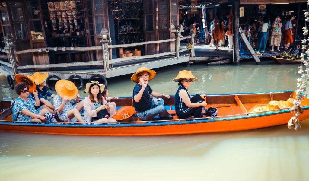 Tourists wearing hats enjoying a guided boat ride through a floating market, with shops and people visible on the wooden platform in the background.