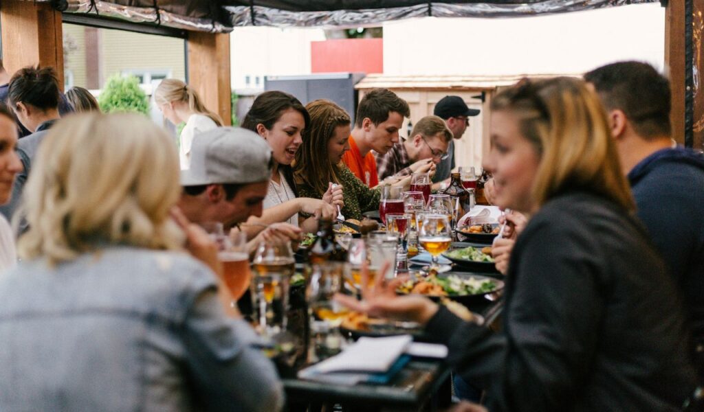 A large group of people enjoying a shared meal at a long outdoor dining table, chatting and sampling a variety of dishes and drinks during a lively food tour experience.