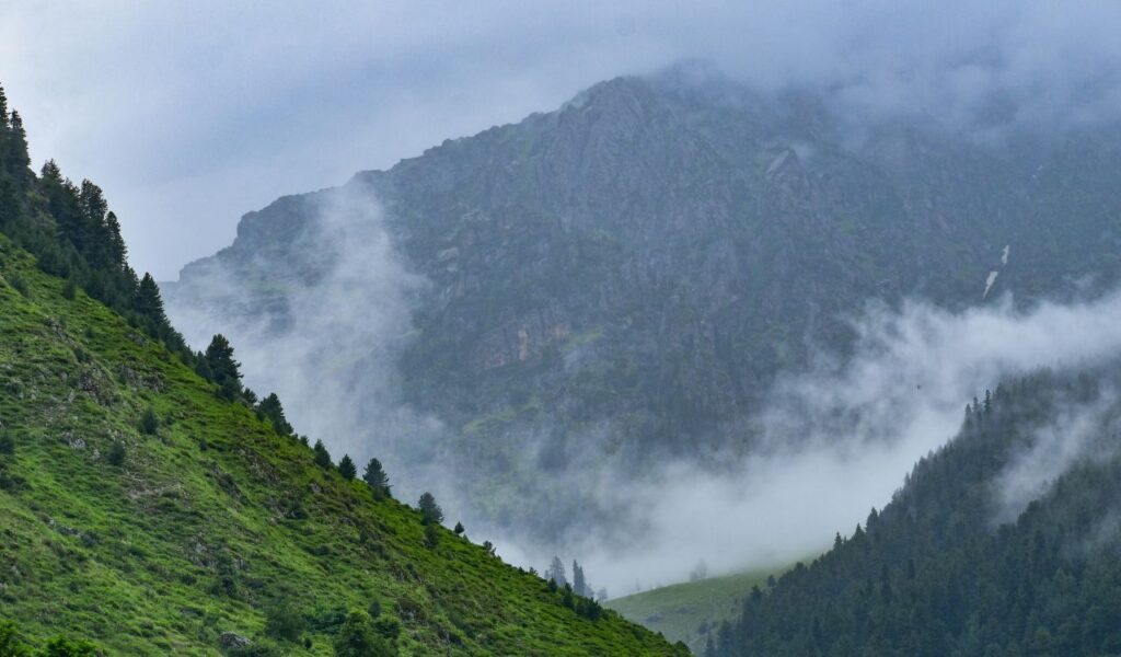 Mist drifts between lush green hills and steep rocky mountains in the foggy highlands of Kashmir, India.