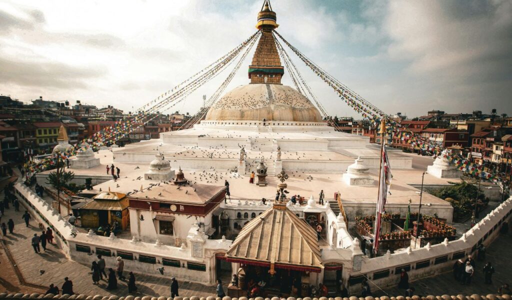 The Boudhanath Stupa in Kathmandu, Nepal, surrounded by prayer flags, pigeons, and people walking around its circular base in a traditional spiritual circuit.