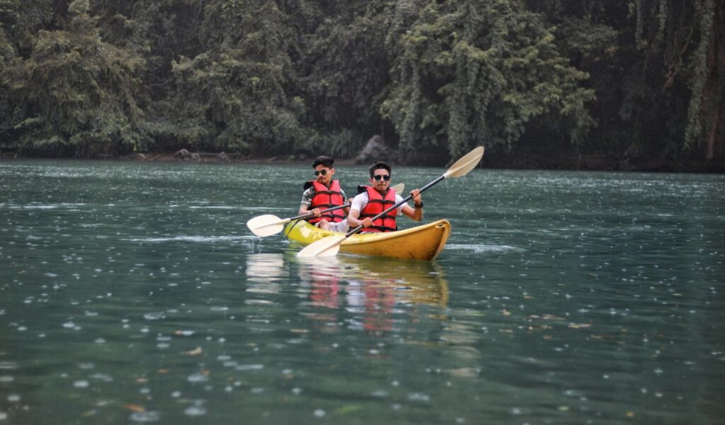 Two men in red life jackets kayaking together on a calm, green river surrounded by dense forest.