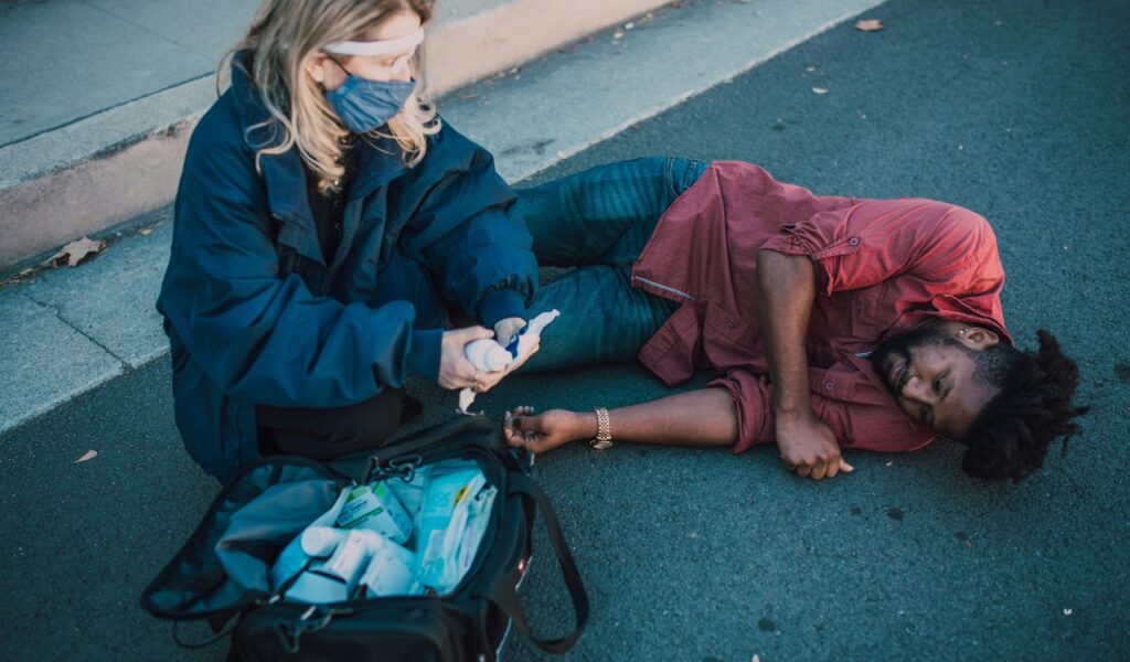 A first responder in a mask tends to a man lying on the pavement, using supplies from a medical emergency kit on the ground beside them.