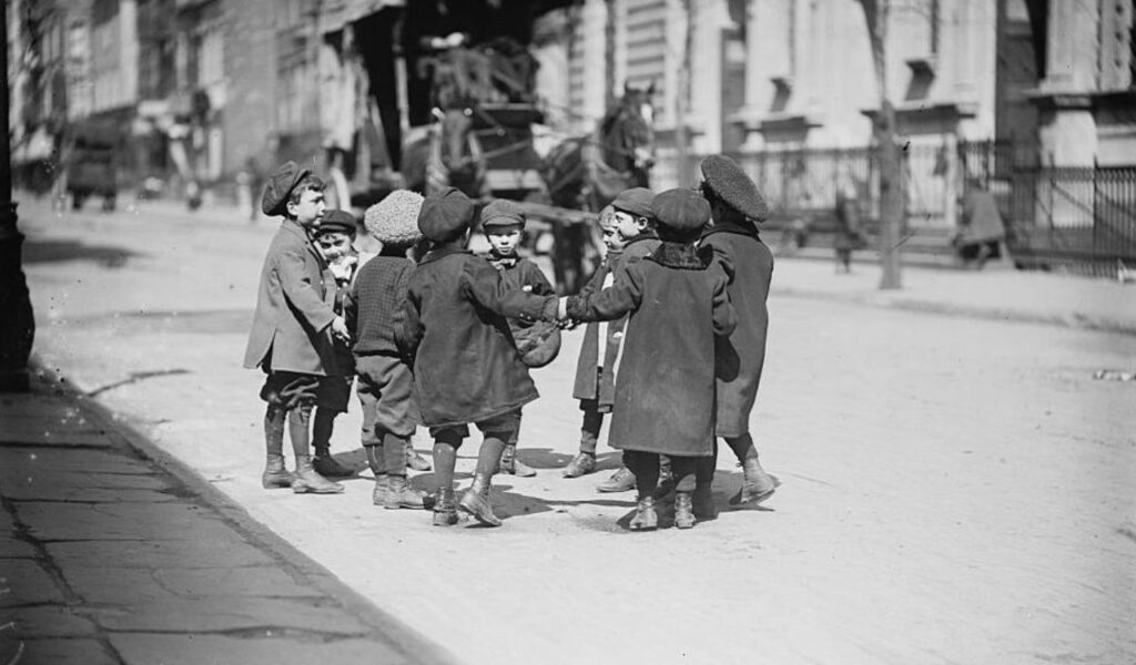 A group of young children hold hands and play in a circle on a city street, dressed in vintage coats and caps.