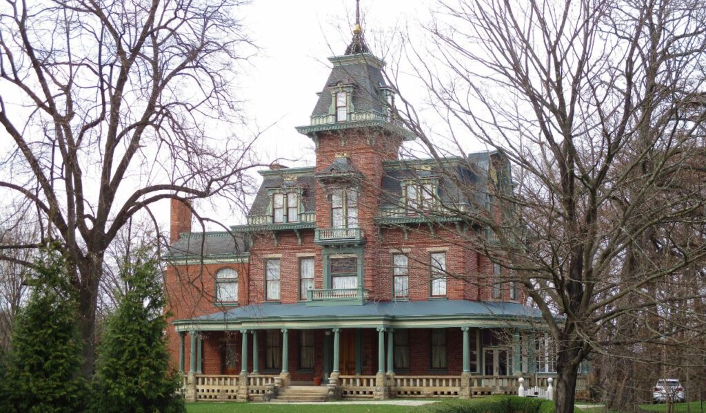 A large, historic brick mansion with ornate architectural details and a wraparound porch, surrounded by leafless trees.