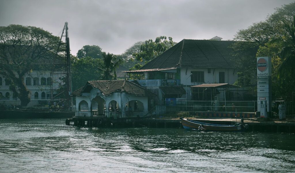 A riverside hotel and Indian Oil petrol station in Kochi, India, with traditional tiled roofs, a small pier, and boats docked near the water under overcast skies.