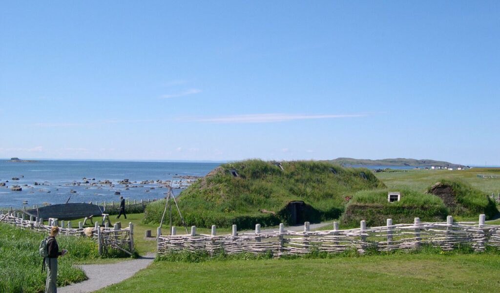 L’Anse aux Meadows, Newfoundland, showing turf-covered Norse-style houses near the rocky shoreline under a clear sky.