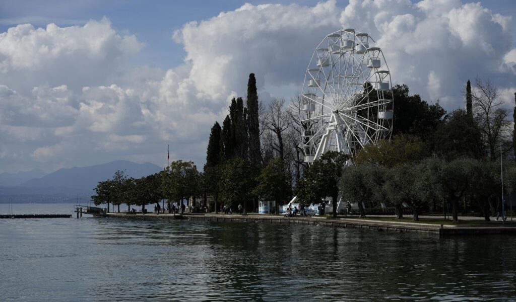 A Ferris wheel near a lakeside surrounded by trees and walking paths under a partly cloudy sky at an amusement park.