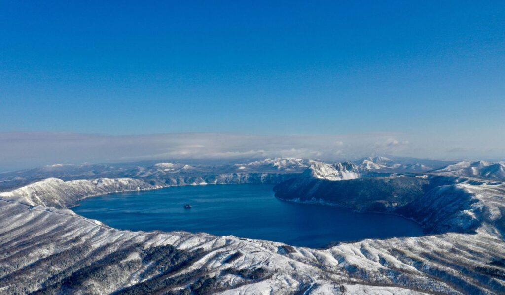 Aerial view of Lake Mashu in Japan, surrounded by snow-covered volcanic mountains under a clear blue sk