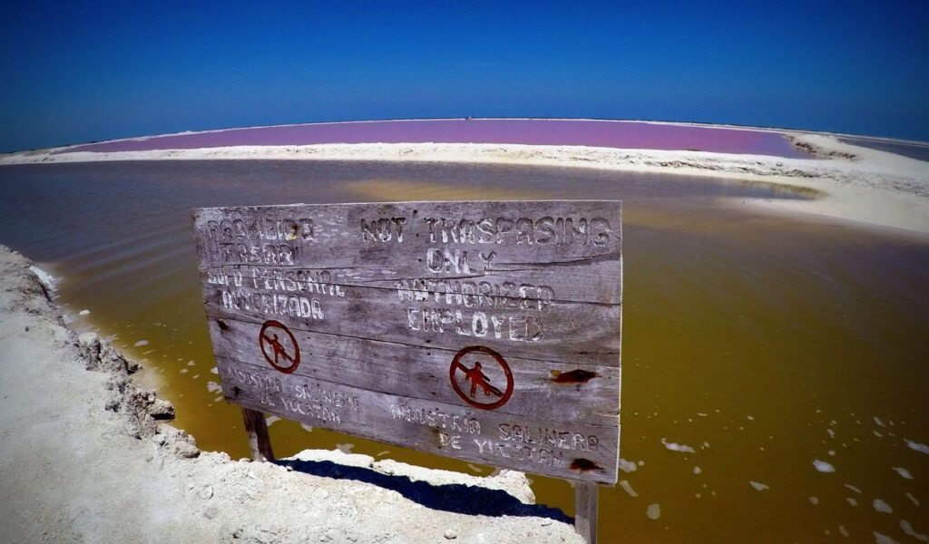 Weathered sign reading “No Trespassing” in front of colorful pink saltwater lagoon at Las Coloradas, Yucatán, Mexico.