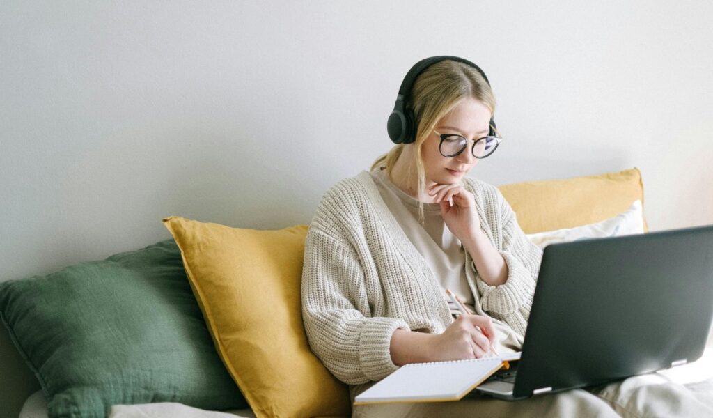Woman wearing headphones and glasses, learning online while taking notes on a notepad, seated on a sofa with colorful cushions.