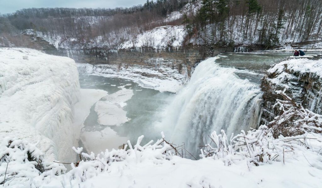 Frozen waterfall cascades into an icy gorge surrounded by snow-covered cliffs and leafless trees in Letchworth State Park.