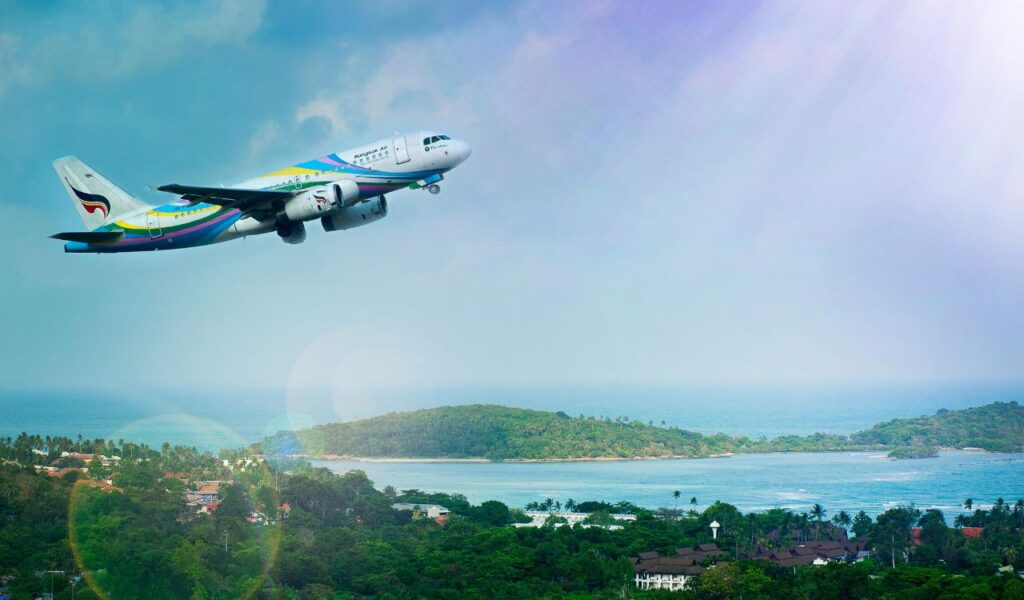 A colorful commercial airplane takes off above a tropical coastline, with blue sky, ocean, and green islands below.
