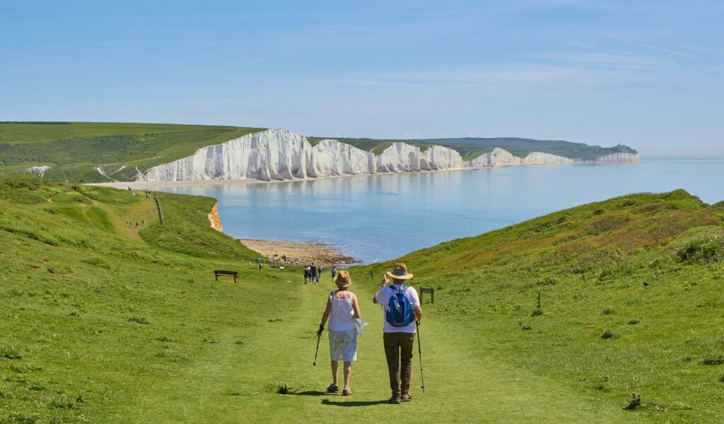 Two older adults walk with trekking poles along a grassy path toward white chalk cliffs and a calm blue sea, enjoying a coastal hike.