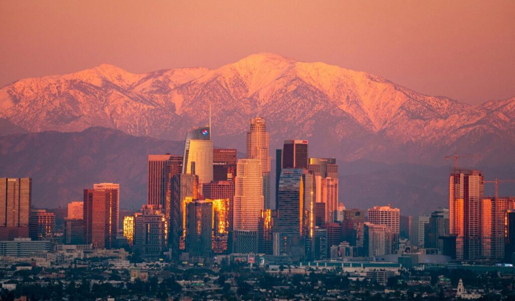 Los Angeles skyline at sunset with snow-capped San Gabriel Mountains in the background, reflecting golden light on city skyscrapers.