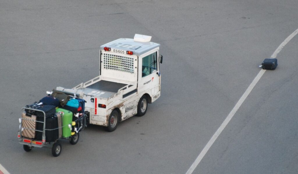 Airport baggage tug hauling packed luggage cart drives past a lone suitcase fallen onto the runway service road.