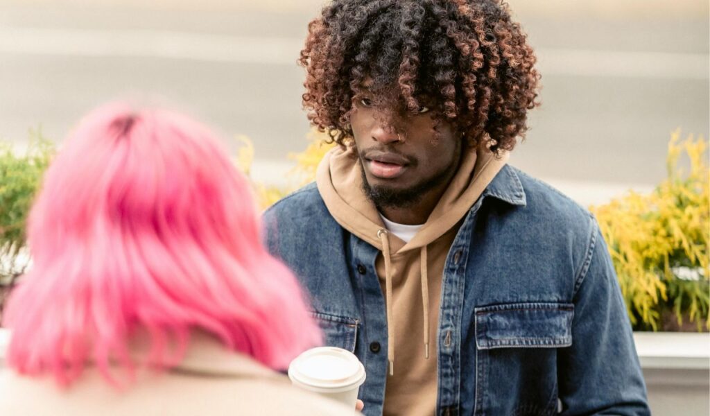 A man listens intently during a conversation at an outdoor café while holding a coffee cup, with another person seated across from him.