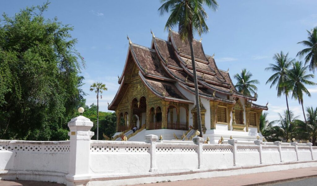Traditional Buddhist temple with ornate gold detailing and a steep, multi-tiered roof in Luang Prabang, Laos, surrounded by palm trees and enclosed by a white fence.