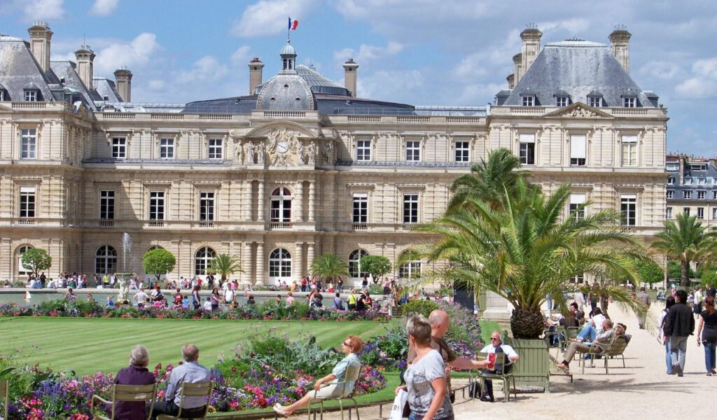 Visitors relax and stroll through the gardens of Luxembourg Palace in Paris, surrounded by colorful flowers and palm trees on a bright sunny day.