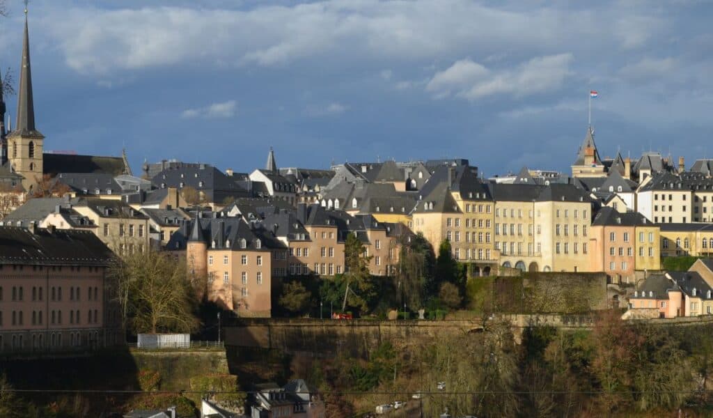Panoramic view of Luxembourg City’s old town with pastel-colored buildings, steep slate rooftops, church spires, and the Luxembourg flag flying above.