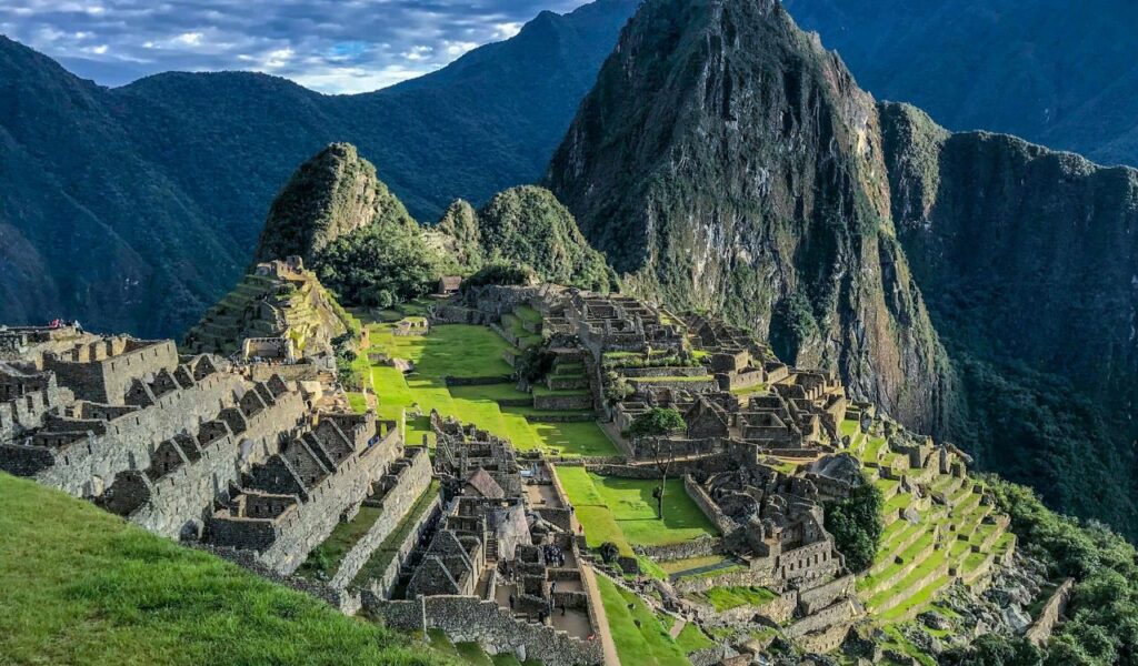Machu Picchu in Peru with stone ruins and terraces set among green mountains under a partly cloudy sky.