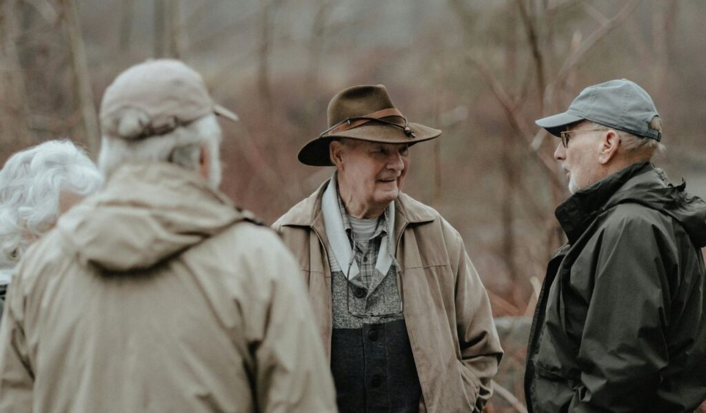 A group of older adults dressed in jackets and hats stand outdoors, chatting and smiling in a relaxed, natural setting.