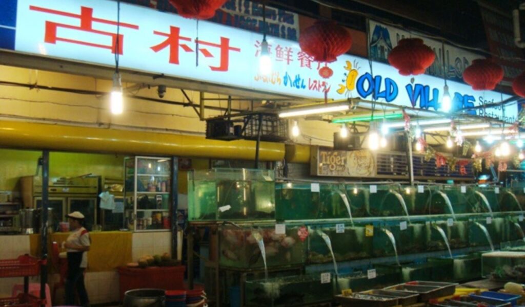 Nighttime view of a seafood street food market in Malaysia, featuring fish tanks, colorful signage, and local food vendors.