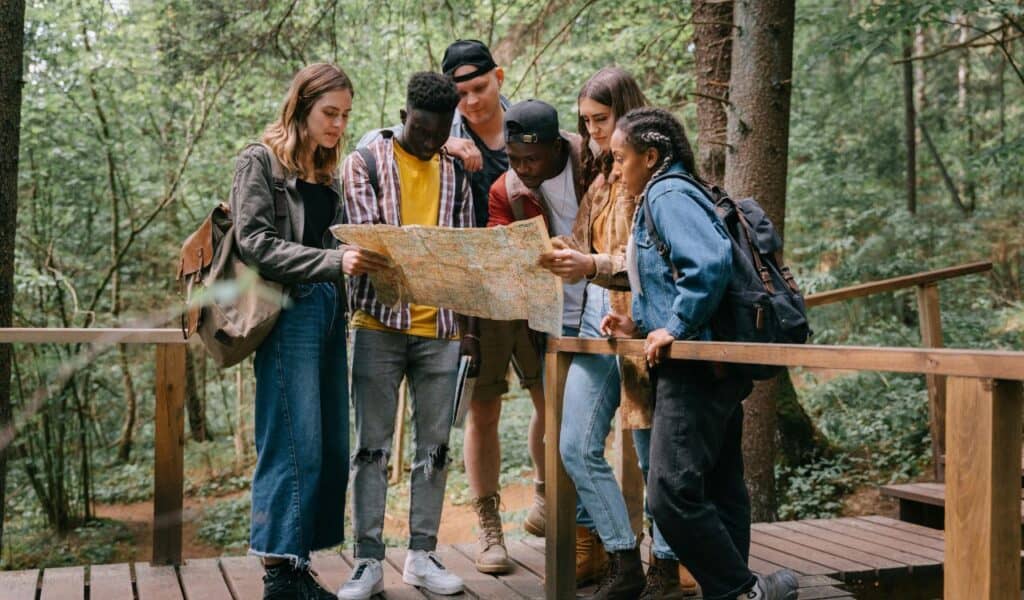 A group of young adults with backpacks stands on a wooden trail bridge in a forest, gathered around a large paper map, planning their route together.