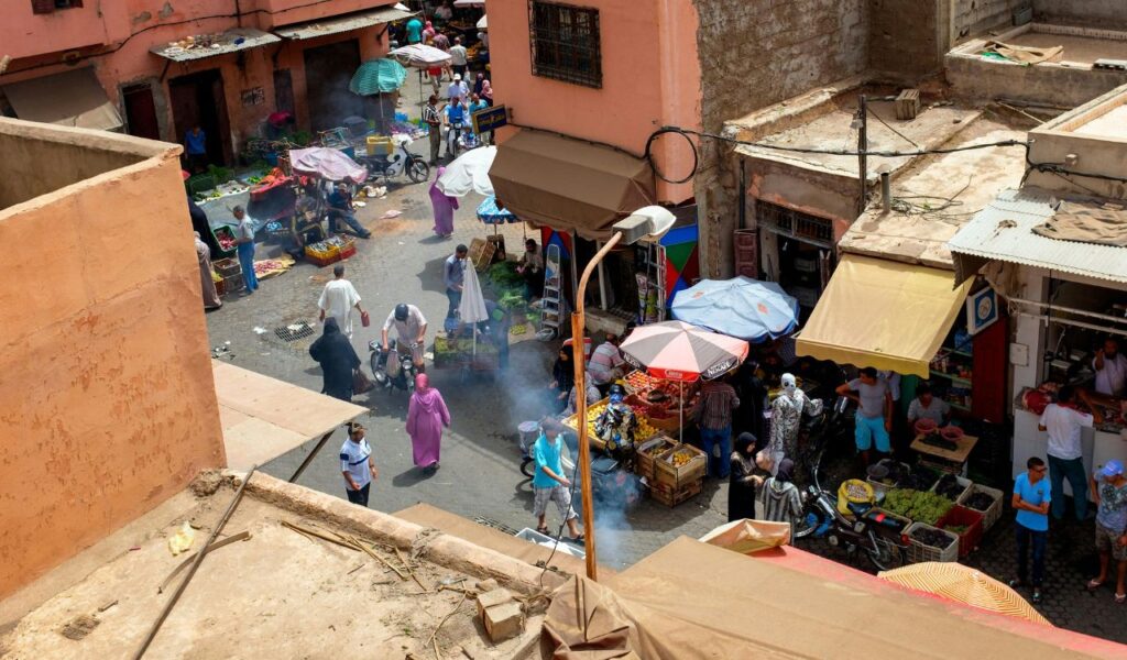 A busy street market in Marrakesh with vendors selling fruits and goods under umbrellas, surrounded by traditional buildings and shoppers.
