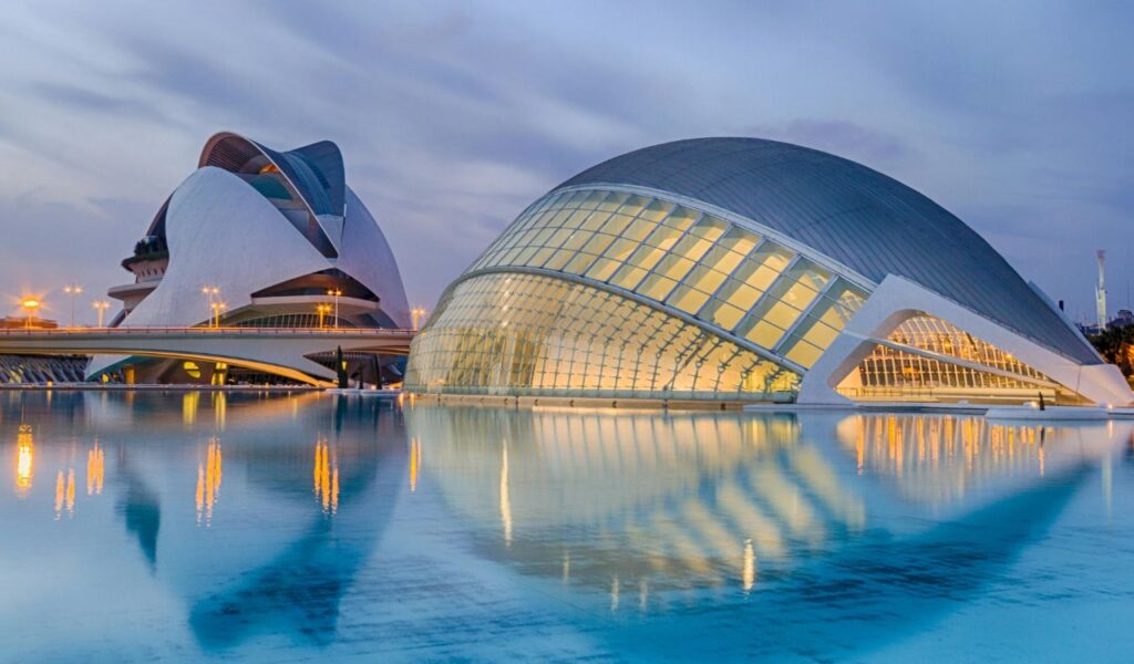 Futuristic buildings with curved glass and metal structures reflected in a shallow blue pool at dusk in a modern architectural complex.