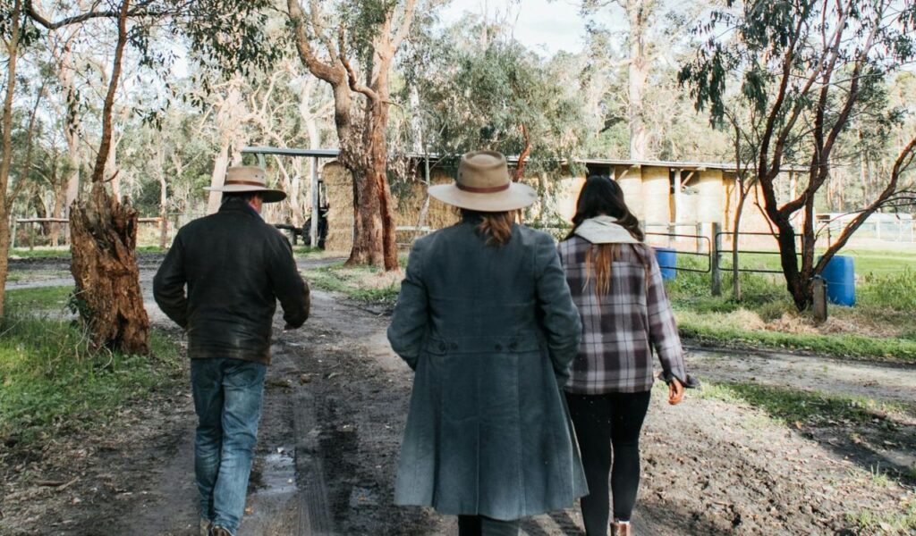 Three people wearing jackets and hats walk along a muddy path toward a rustic farm surrounded by trees and hay bales.