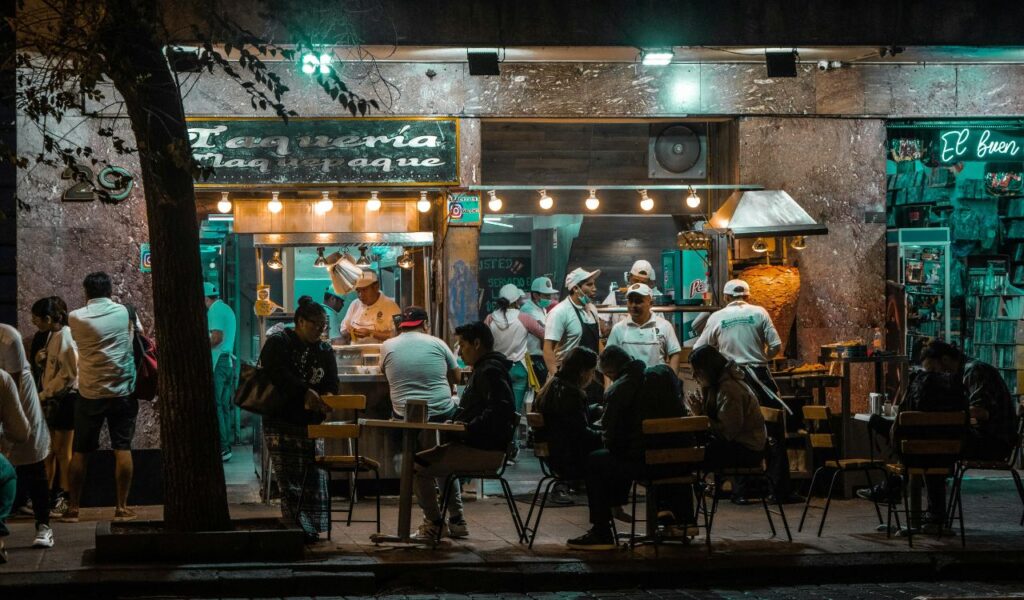Nighttime street food scene in Mexico City with people dining at outdoor tables while chefs prepare tacos inside a taqueria.