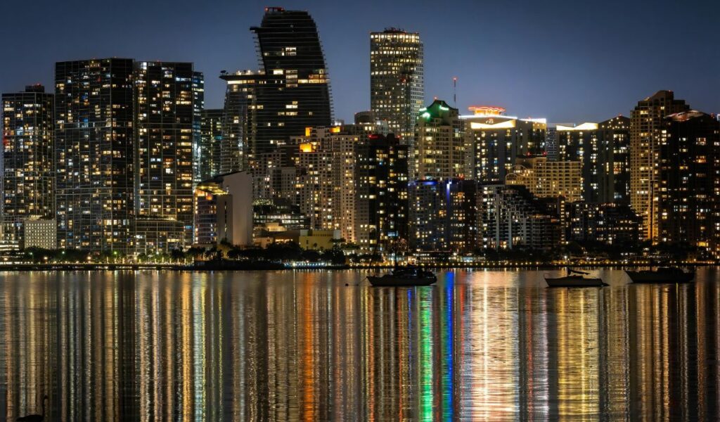 Night view of Miami skyline with illuminated buildings reflecting on the water and boats anchored in the foreground