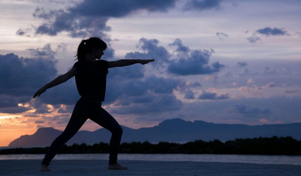 Silhouette of a person practicing yoga at sunset beside water, with distant mountains and dramatic clouds.