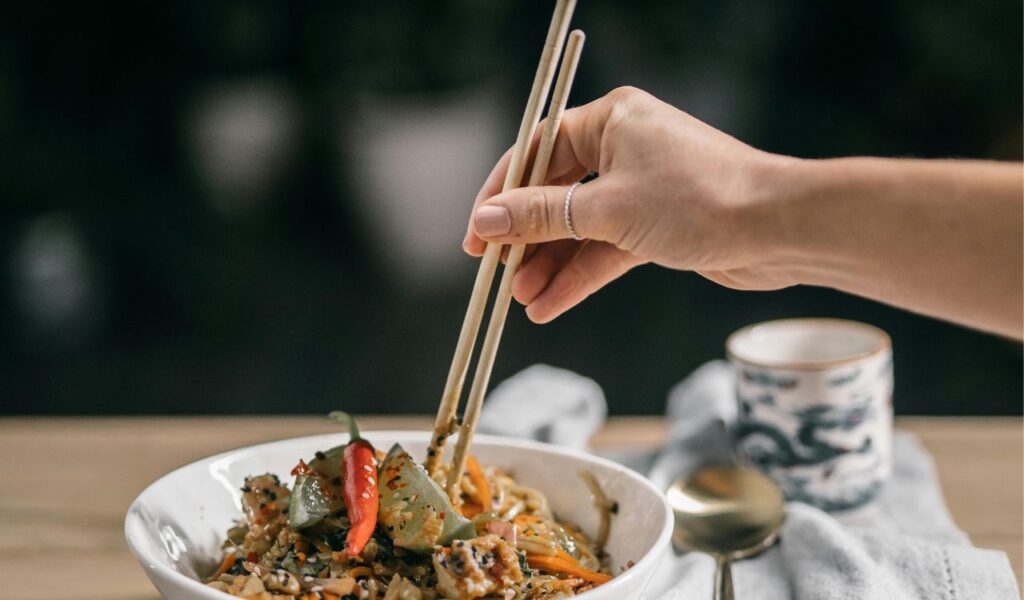 Hand holding wooden chopsticks picking up food from a bowl of noodles and vegetables, with a ceramic tea cup and spoon in the background.