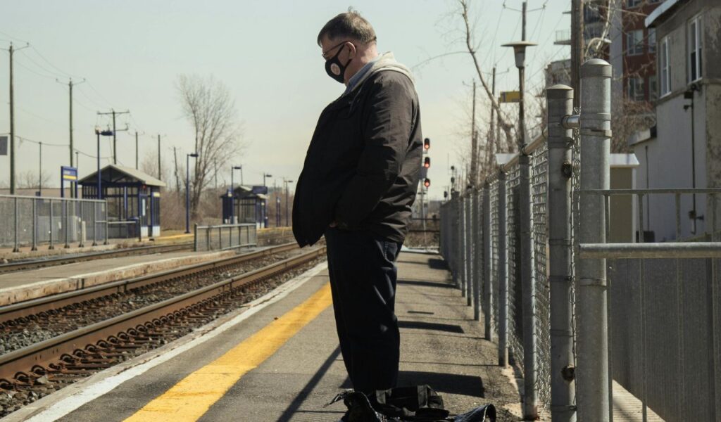 A man wearing a mask stands alone on a train platform next to railway tracks, waiting for the train on a quiet, empty station.