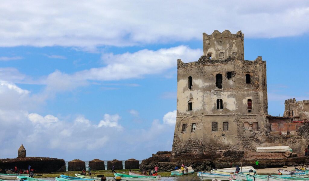 A crumbling historic tower stands along the waterfront in Mogadishu, Somalia, surrounded by small wooden fishing boats and people on the shore, under a partly cloudy sky.