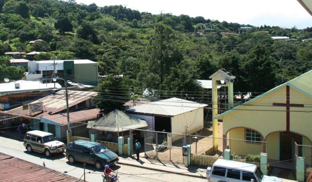 Street view of Monteverde, Costa Rica, showing local buildings, vehicles, and lush green forested hills in the background.