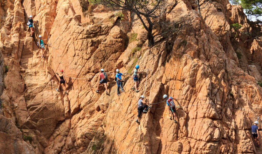 A group of climbers wearing helmets and harnesses ascend a steep rocky cliff using a Via Ferrata route with fixed cables and ladders.