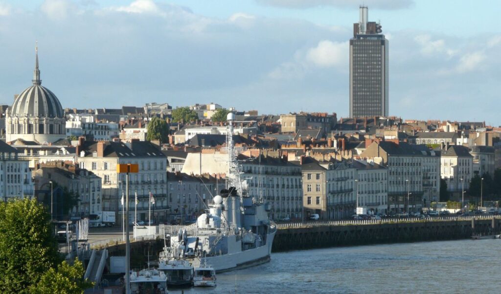 Nantes cityscape along the Loire River, featuring classic French buildings, a modern tower, and a docked naval ship in the foreground.