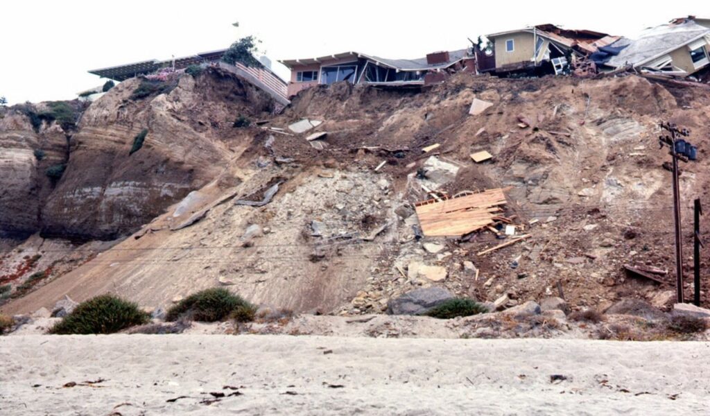 Clifftop houses wrecked and sliding after a landslide, debris and earth cascading down the exposed slope.