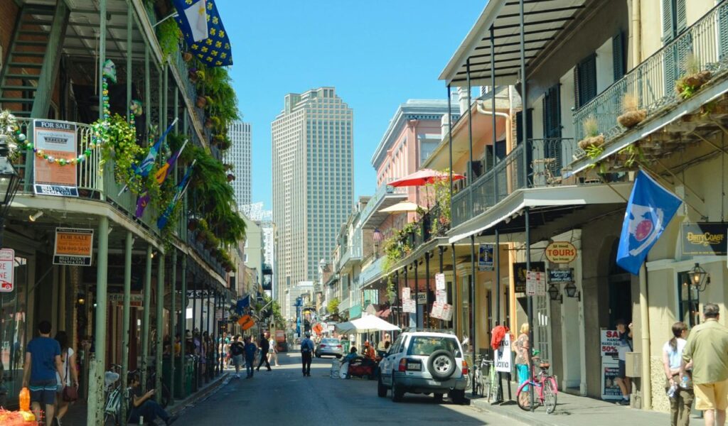 Street view in New Orleans French Quarter with historic buildings, hanging plants, flags, pedestrians, and modern skyscrapers in the background