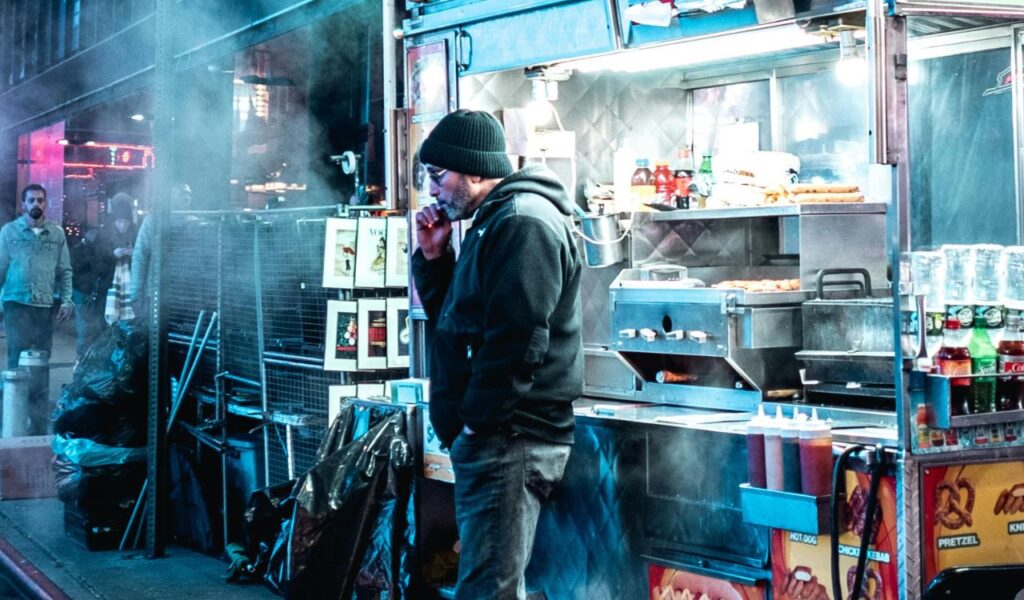 A man standing by a steaming food cart in New Orleans at night, with hot dogs, pretzels, and condiments visible in the brightly lit stall.
