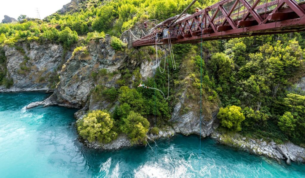 A person bungee jumps off a red suspension bridge above a turquoise river, surrounded by rocky cliffs and lush green trees.