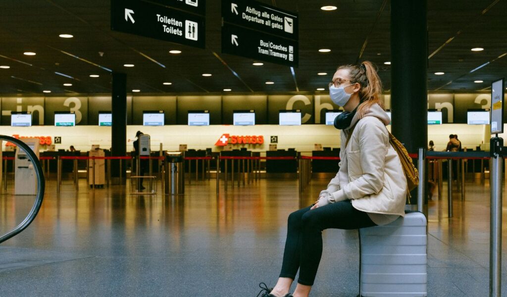 A masked traveler sits on a suitcase in an airport terminal near the SWISS check-in area under departure signs.