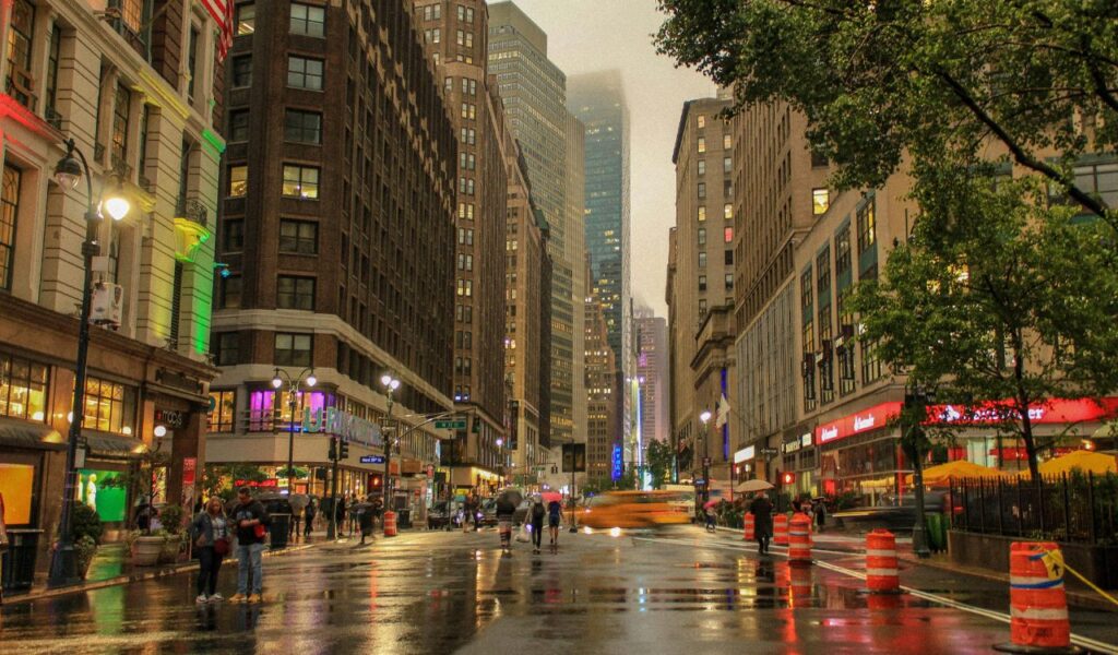 Rain-soaked street in New York City with pedestrians, taxis, and tall buildings under a misty skyline at dusk
